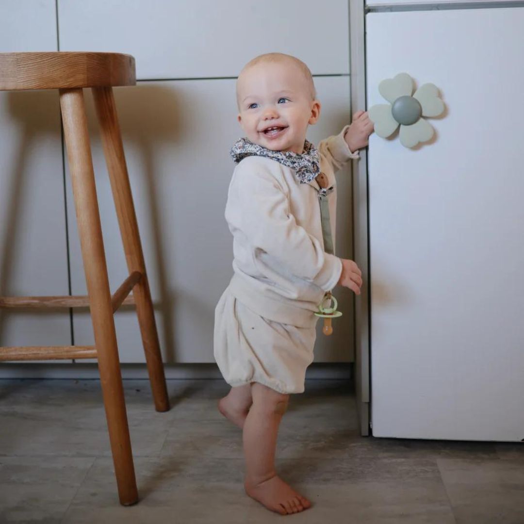 Een lachende baby speelt met de mushie babyspeelgoed spinner in de vorm van een bloem op een witte kastdeur.
