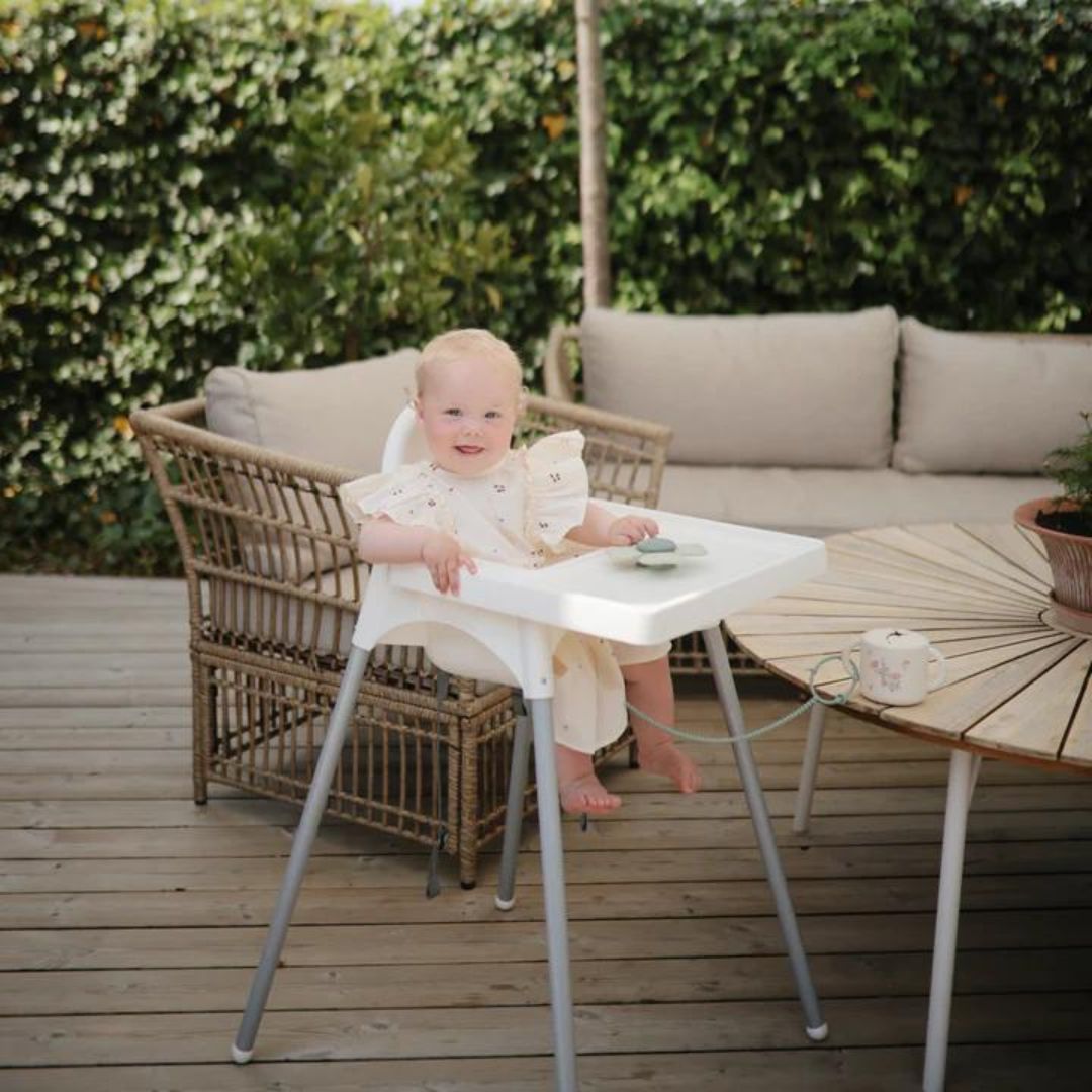 Een baby zit in een kinderstoel buiten en speelt met de mushie babyspeelgoed spinner op het eetblad.