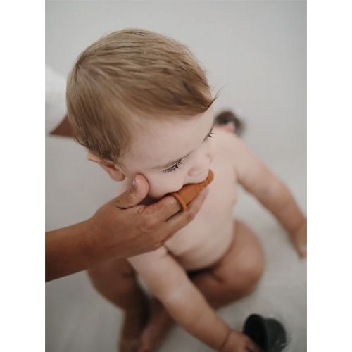 Een volwassen hand gebruikt een Mushie siliconen babytandenborstel bij een baby in bad.