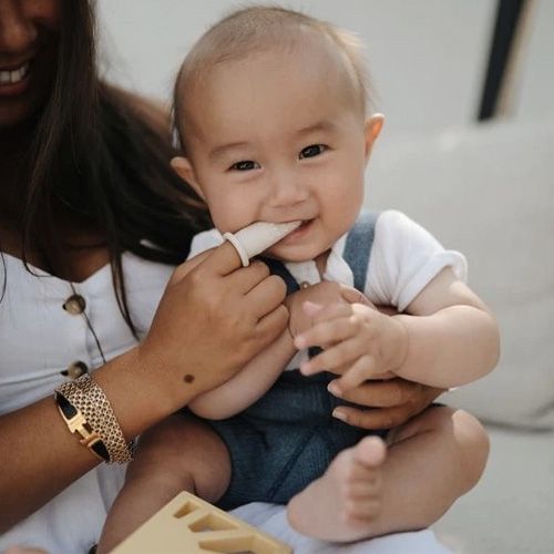 Een lachende baby kauwt op een mushie siliconen babytandenborstel, vastgehouden door een volwassene.