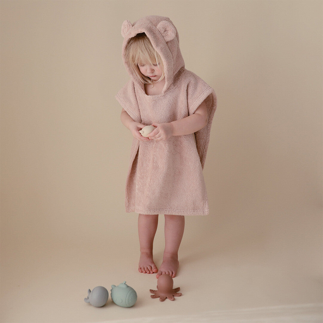 Een peuter draagt een mushie blush roze badponcho met berenoren en speelt met badspeelgoed.