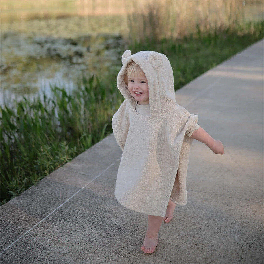 Een lachende peuter draagt de mushie beige badponcho met berenoortjes en loopt blootsvoets buiten.