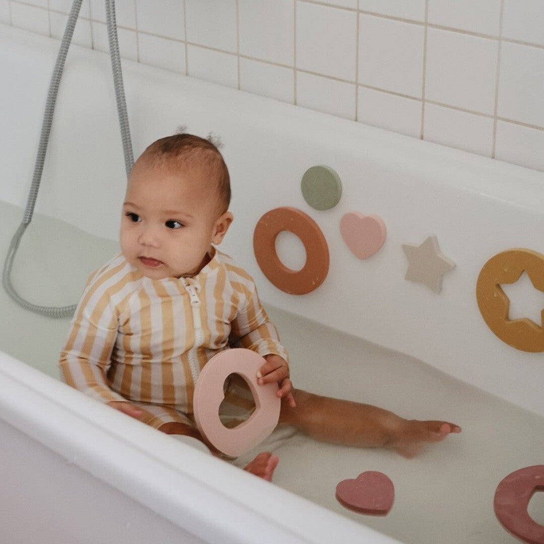 Een baby in een badkuip speelt met mushie badpuzzel foam stukken in pastelkleuren.