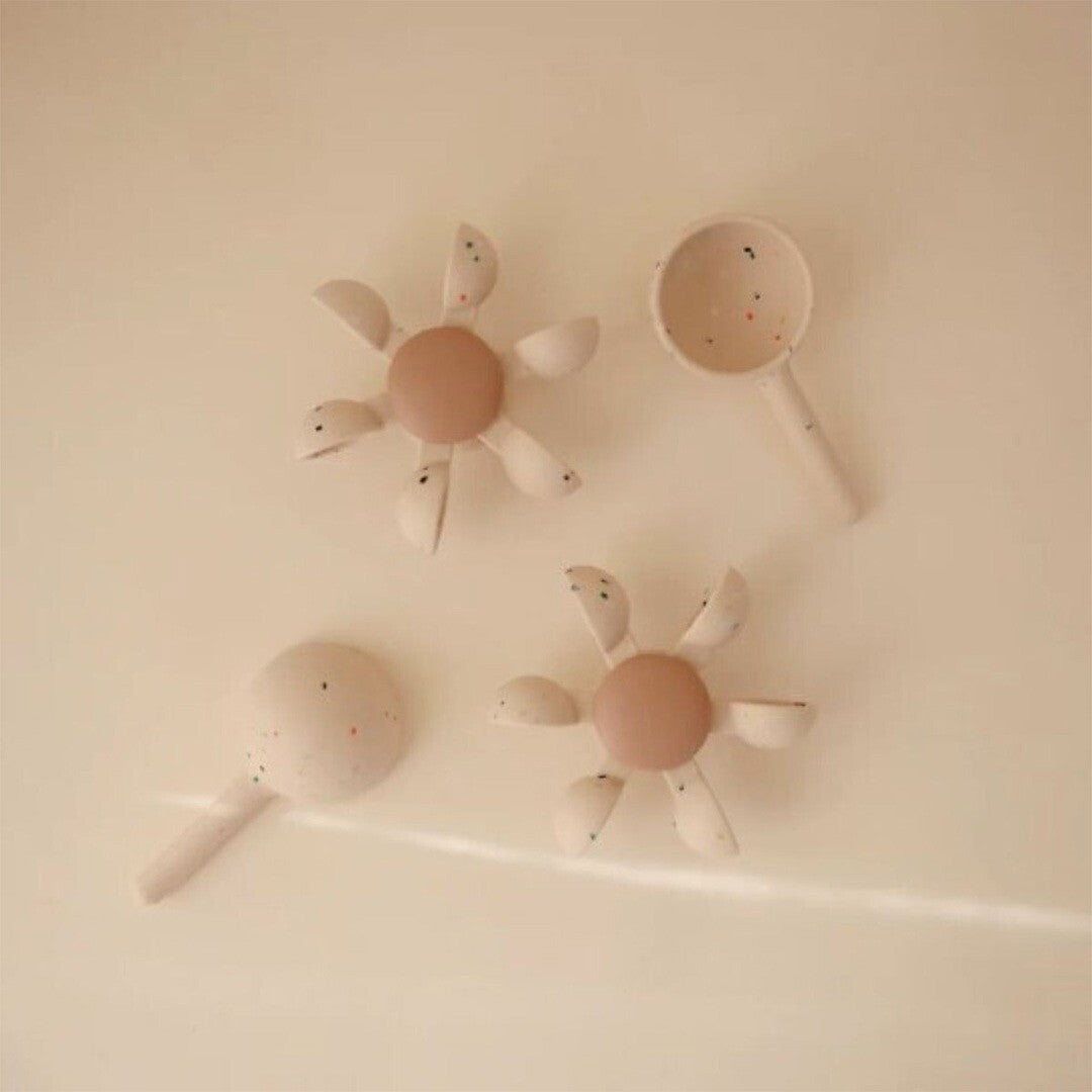 Twee mushie badspeelgoed waterwielen en twee schepjes in beige met kleurrijke spikkels op een lichte ondergrond.