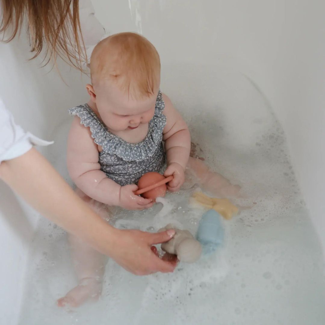 Een baby in bad speelt met de mushie ruimte badspeeltjes, terwijl een volwassene helpt.