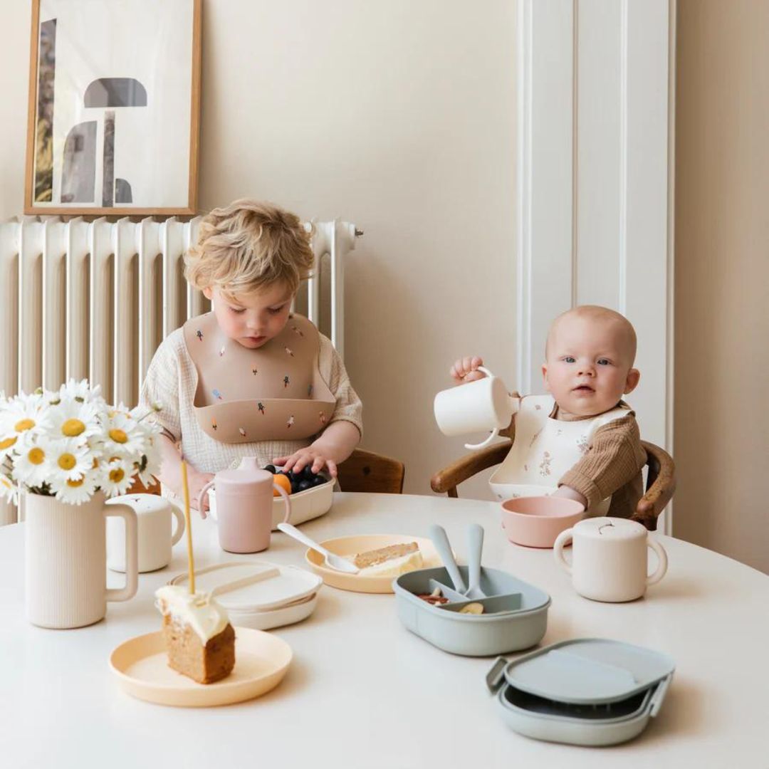 Twee kinderen aan tafel met diverse mushie baby- en peuterservies, waaronder bekers en bordjes.