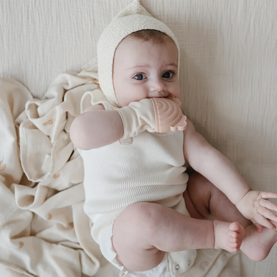 Een baby met een mushie bijthandschoen in de mond, liggend op een lichte deken.