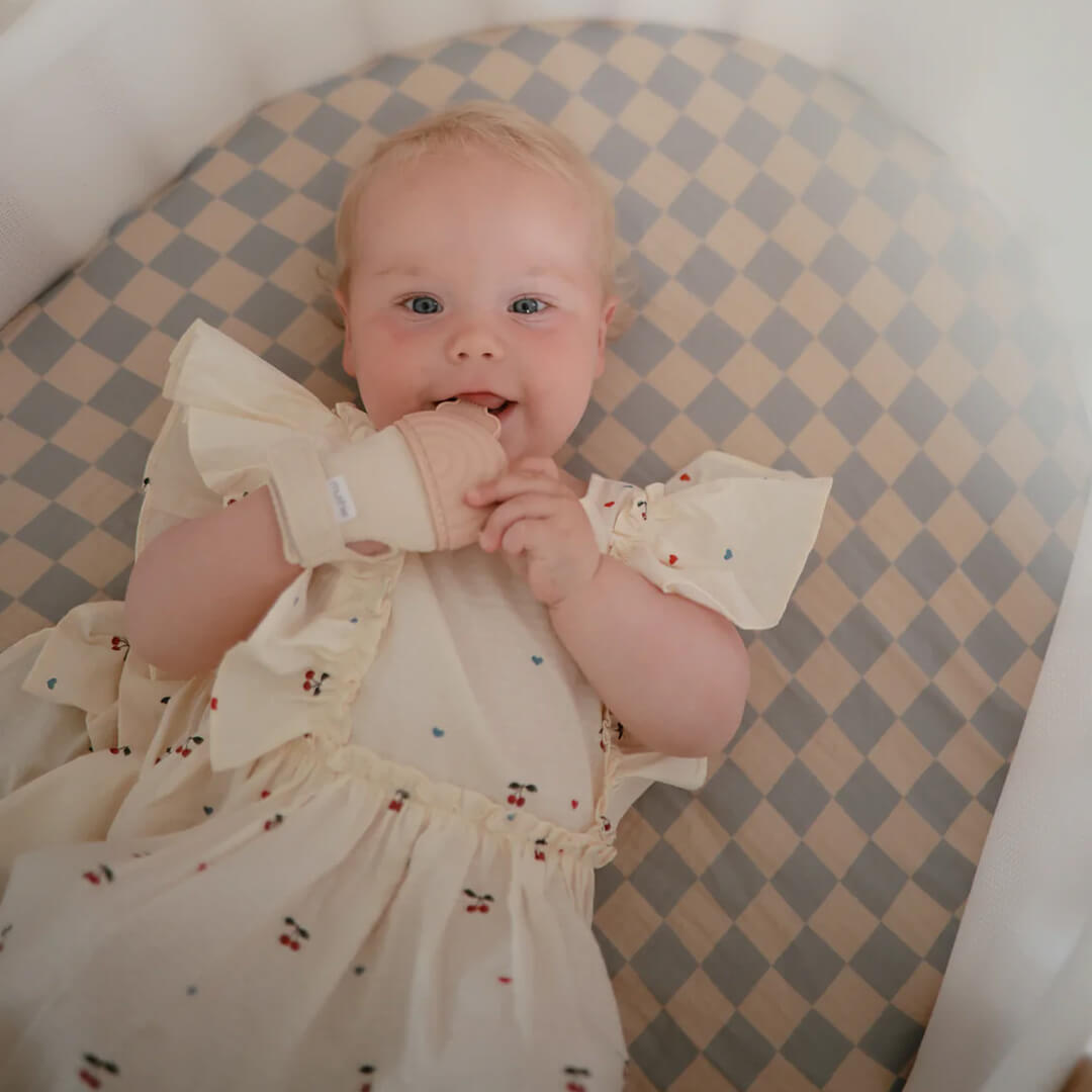 Een lachende baby in een wiegje die op een mushie bijthandschoen kauwt.