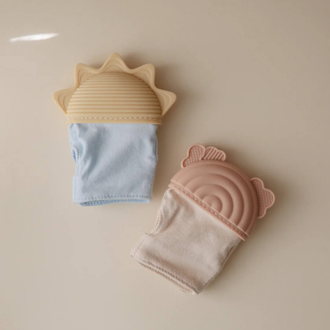 Twee mushie bijthandschoenen, één roze met regenboog en één blauw met zon, liggen naast elkaar.