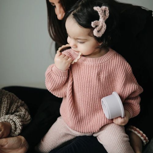 Een baby in een roze trui bijt op een mushie bijtring armband.