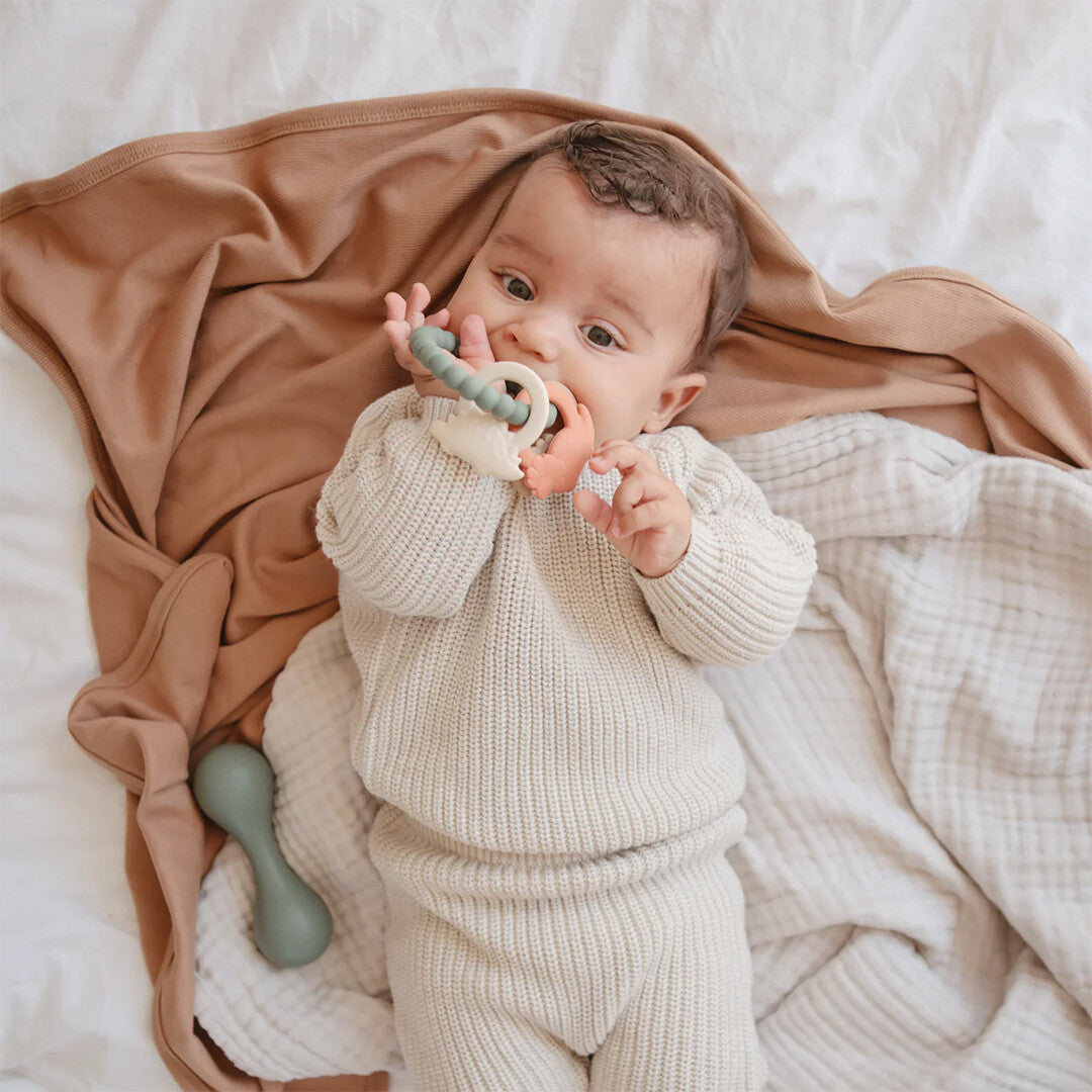 Een baby ligt op een bed en bijt op de mushie dinosaurus bijtring.