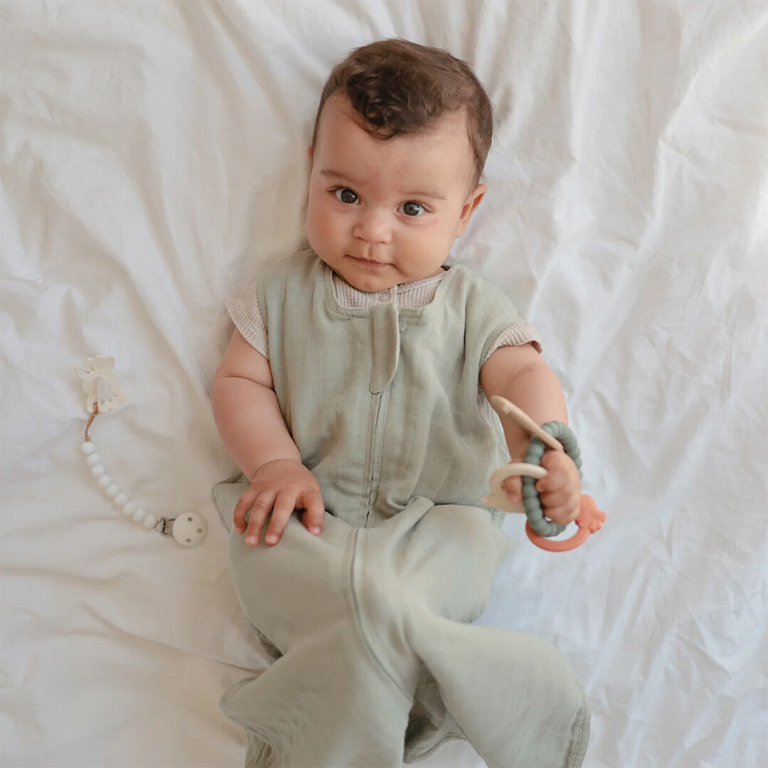 Een baby in een slaapzak houdt de mushie dinosaurus bijtring vast.