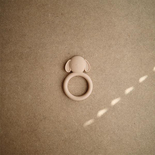 Mushie beige siliconen bijtring in de vorm van een hondenkop, van bovenaf gefotografeerd op een bruine ondergrond.