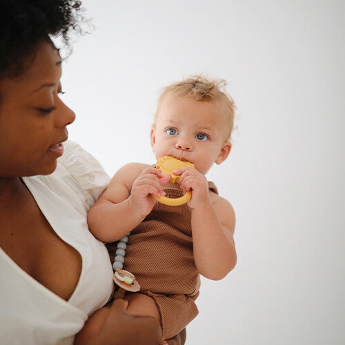 Een baby met blauwe ogen kauwt op de Mushie gele leeuwen bijtring, vastgehouden door een volwassene.