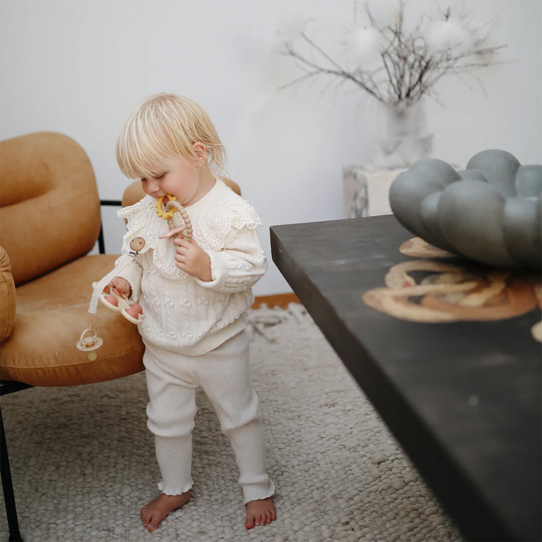 Een blond kindje in een witte gebreide outfit speelt met de Mushie bijtring.