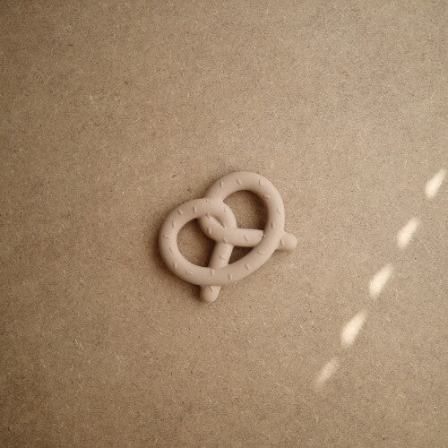 De mushie siliconen bijtring in de vorm van een beige pretzel ligt op een bruine ondergrond.
