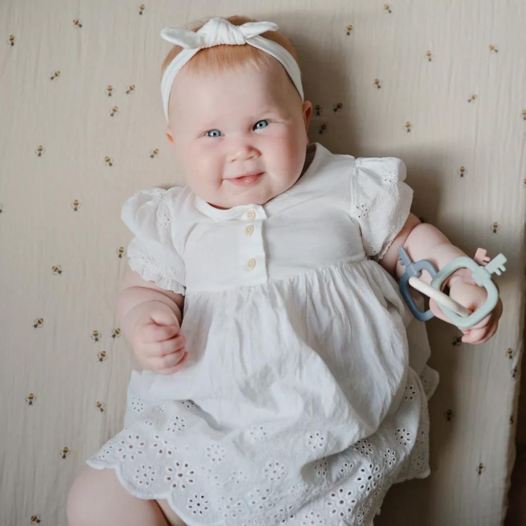 Lachende baby in een witte jurk met een mushie bijtring sleutelbos in de hand.
