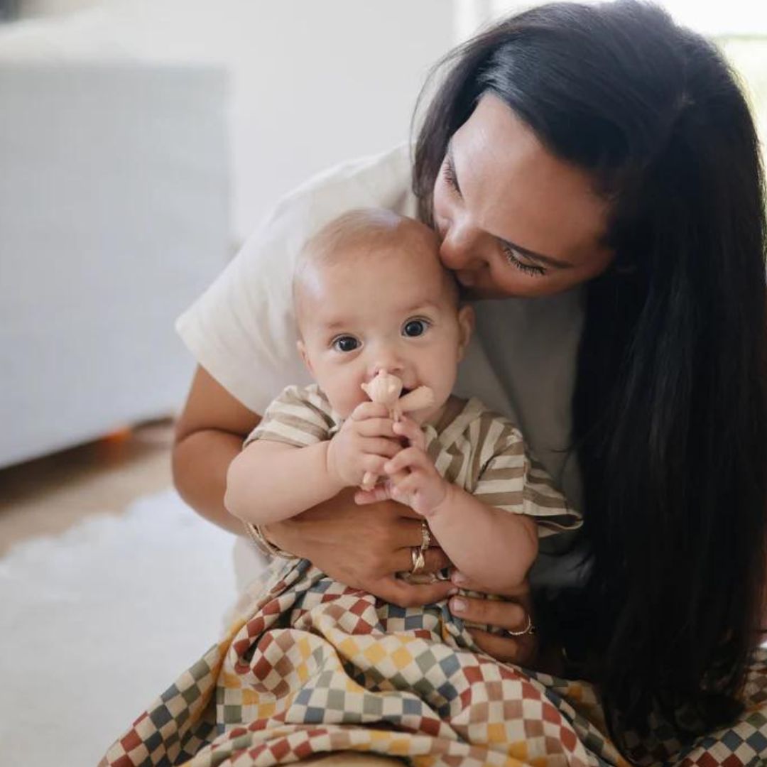 Een baby kauwt op het Mushie roze eenhoorn bijtspeelgoed terwijl een volwassene de baby kust.