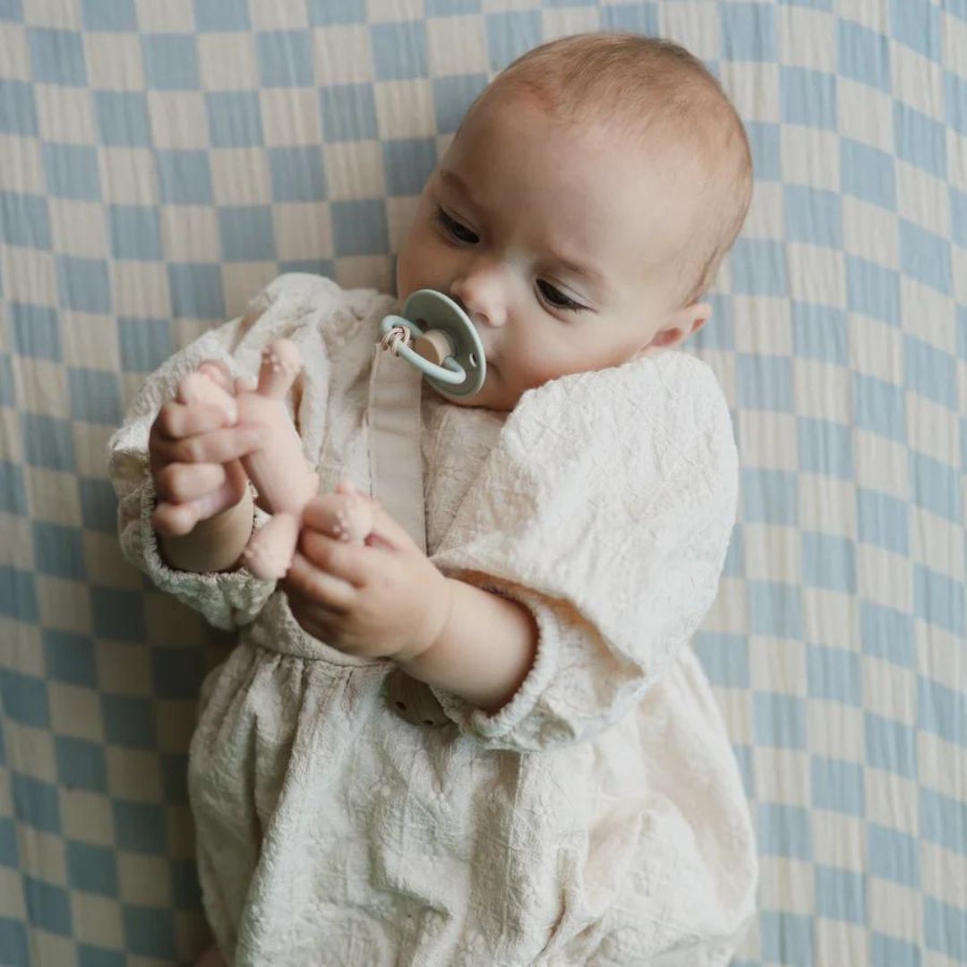 Een baby ligt op een blauw-wit geruite deken en speelt met het Mushie roze eenhoorn bijtspeelgoed.
