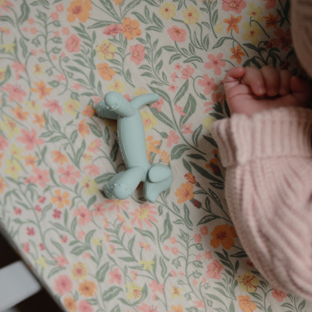 Groene mushie bijtspeelgoed hond naast een babyhand op een bloemenlaken.