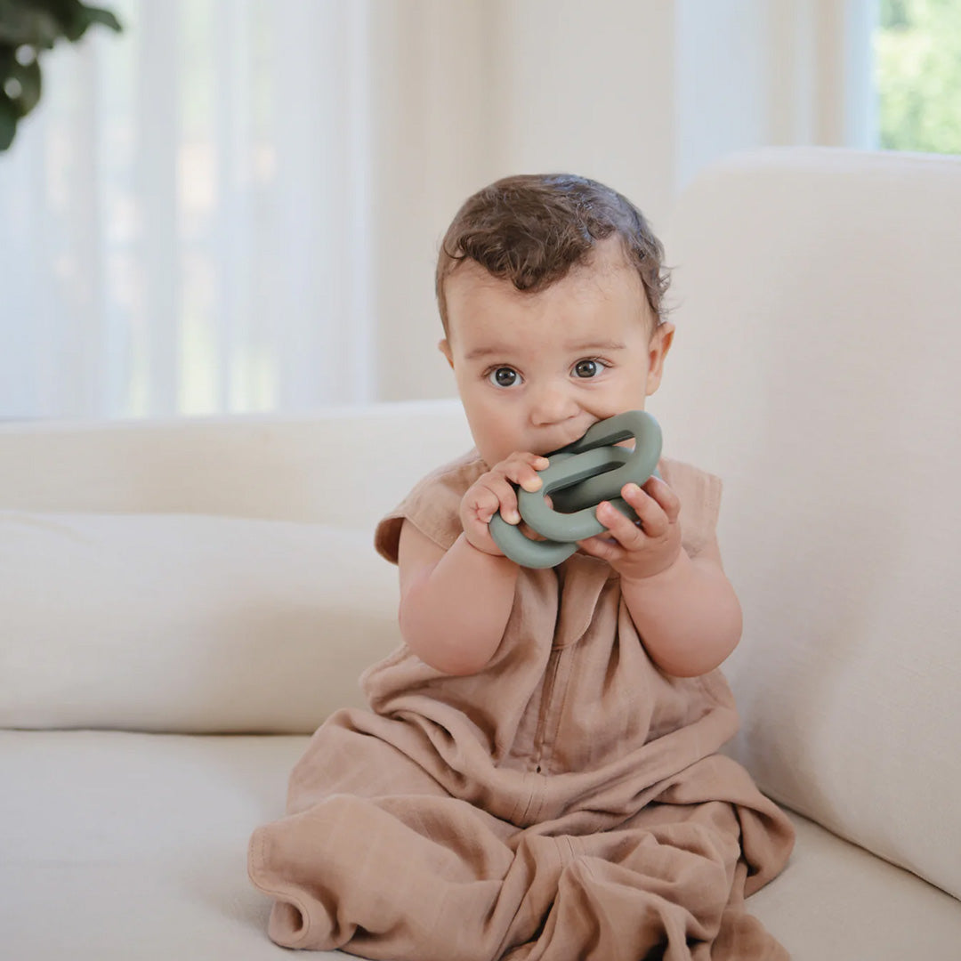 Een baby bijt op het groene mushie bijtspeelgoed zittend op een bank.