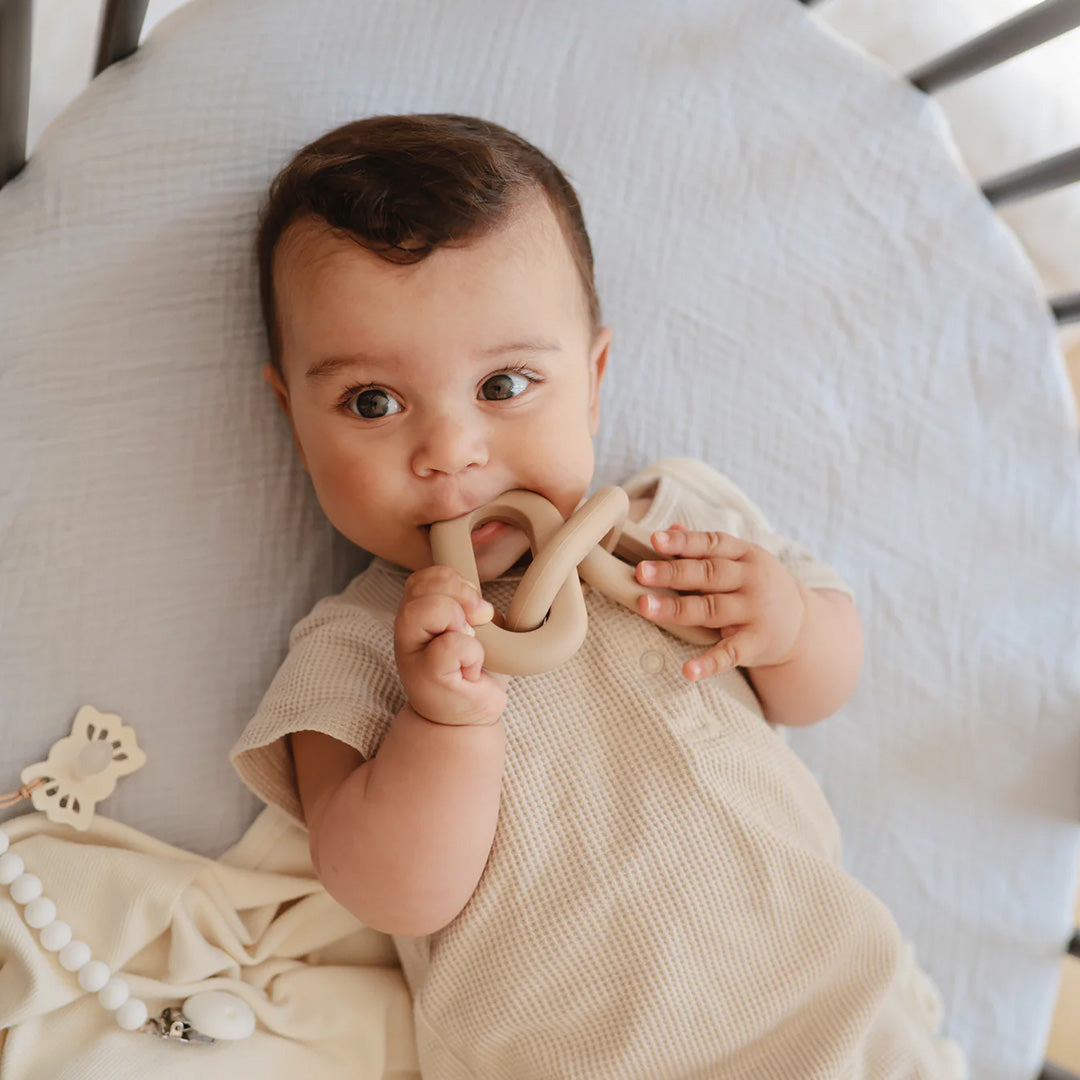 Een baby ligt in een wieg en bijt op een beige mushie bijtspeelgoed schakel.