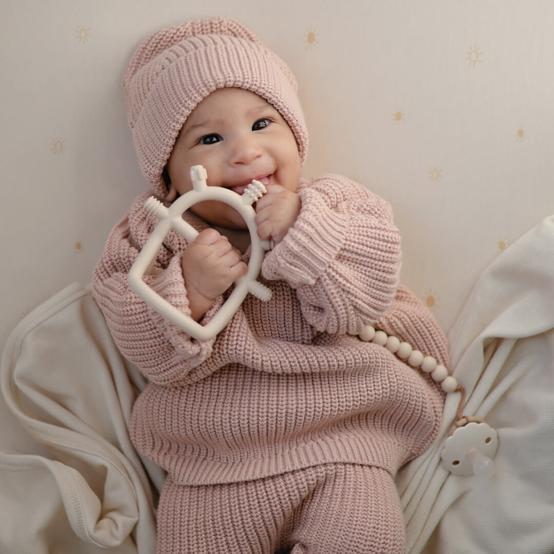 Een baby in een roze gebreid pakje lacht terwijl het op het mushie beige bijtspeelgoed kauwt.