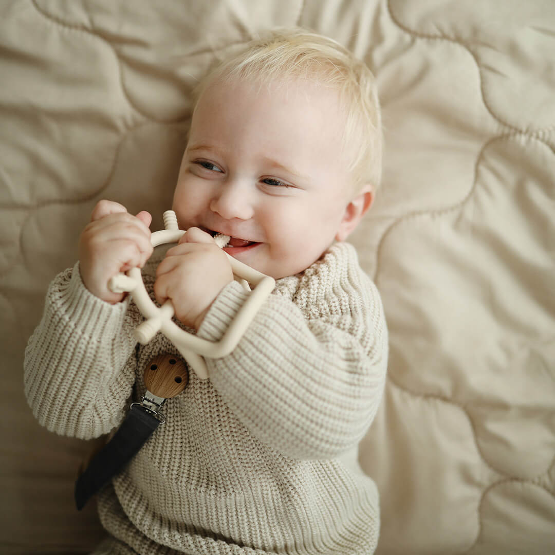 Een lachende baby met blond haar kauwt op het mushie beige bijtspeelgoed.