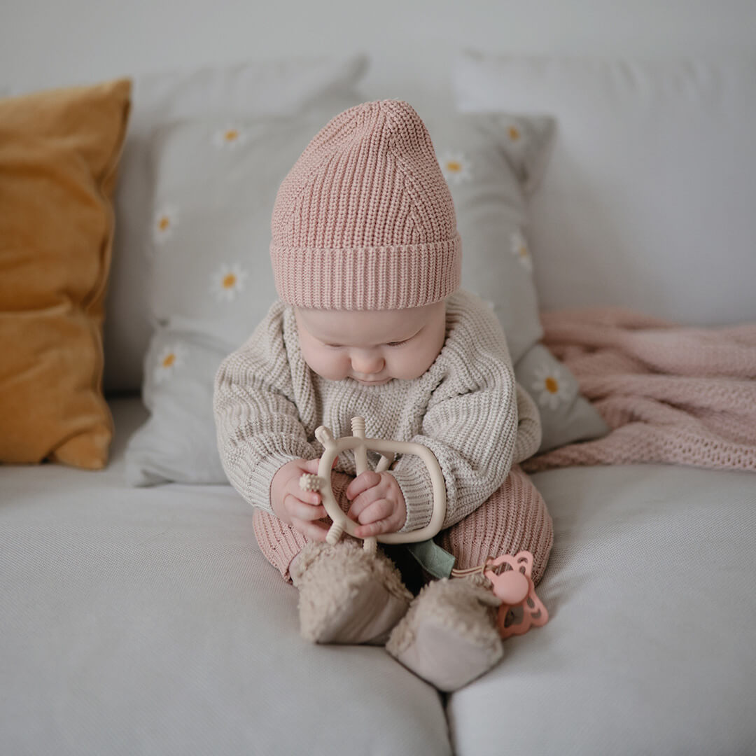 Een baby met een roze muts zit op de bank en houdt het mushie beige bijtspeelgoed vast.