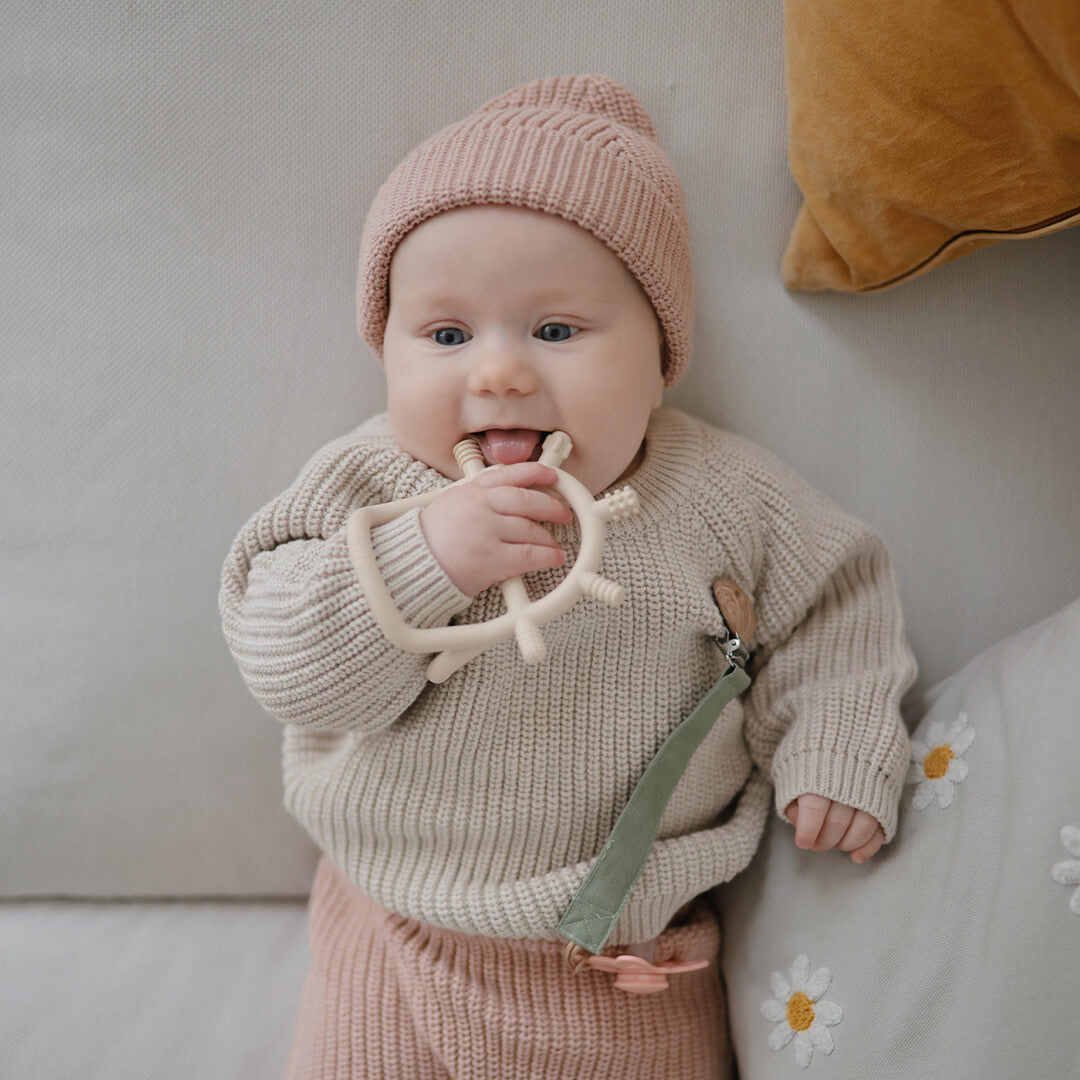 Een baby met een roze muts ligt op de bank en kauwt op het mushie beige bijtspeelgoed.