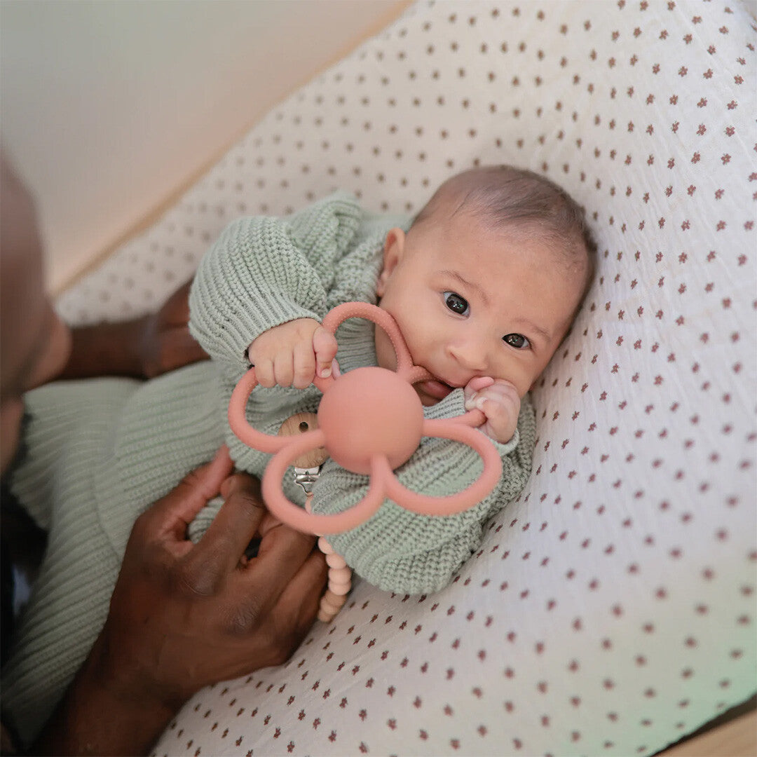 Een baby in een baby nestje houdt het roze mushie bijtspeelgoed in de vorm van een bloem vast.