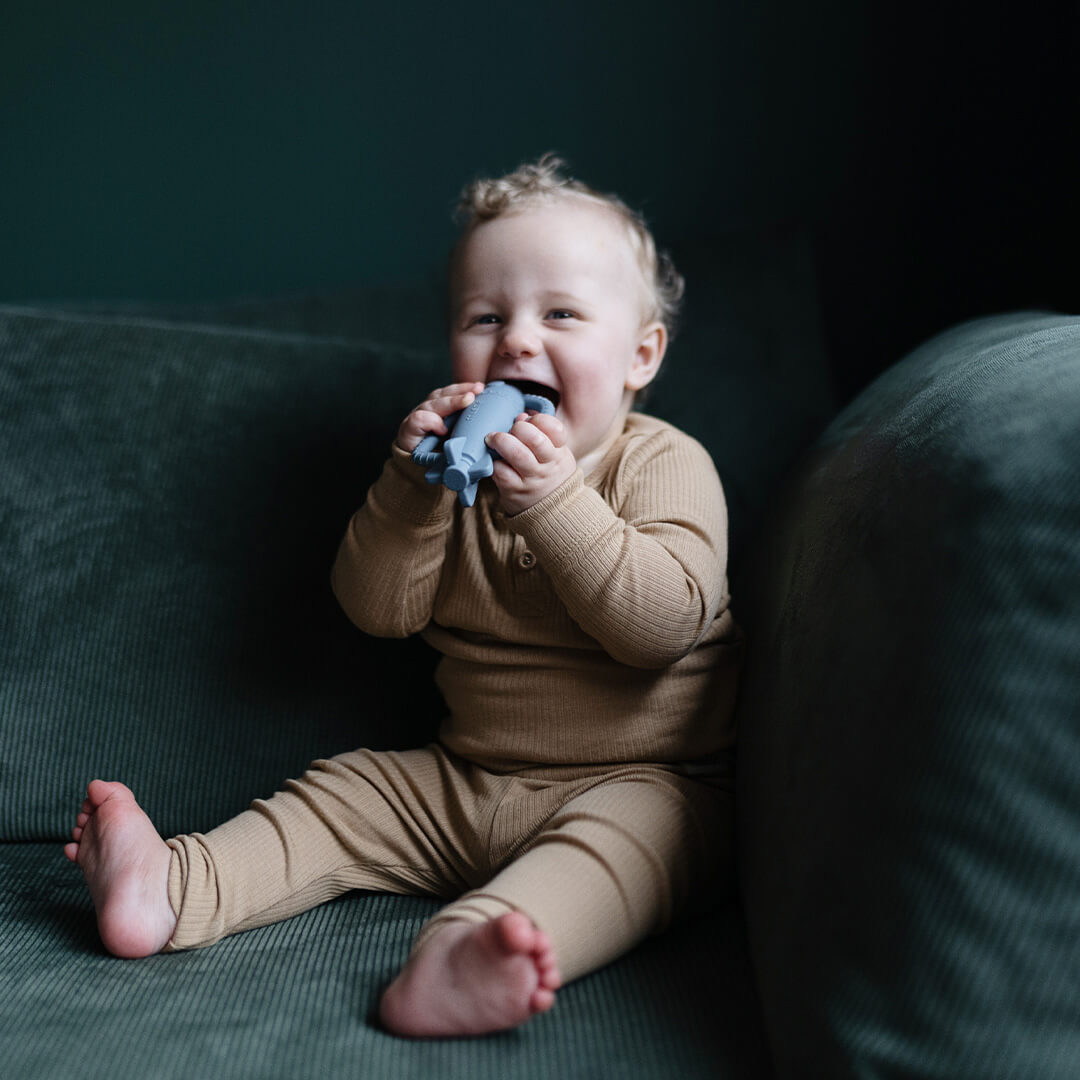 Een lachende baby zit op een groene bank en kauwt op het blauwe raket bijtspeelgoed.