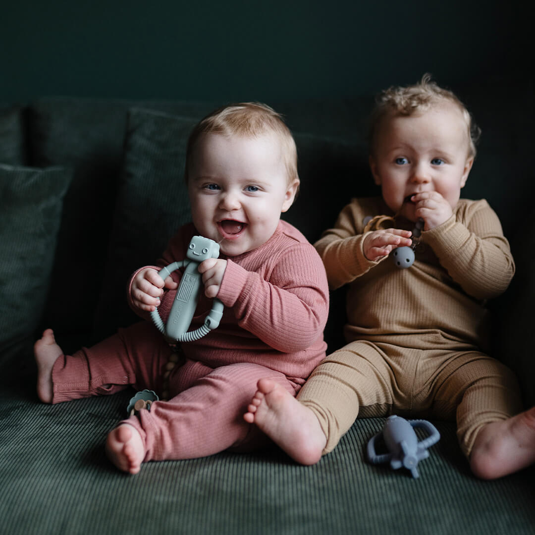 Twee lachende baby's op een groene bank, waarvan één een mushie robot bijtspeelgoed vasthoudt.
