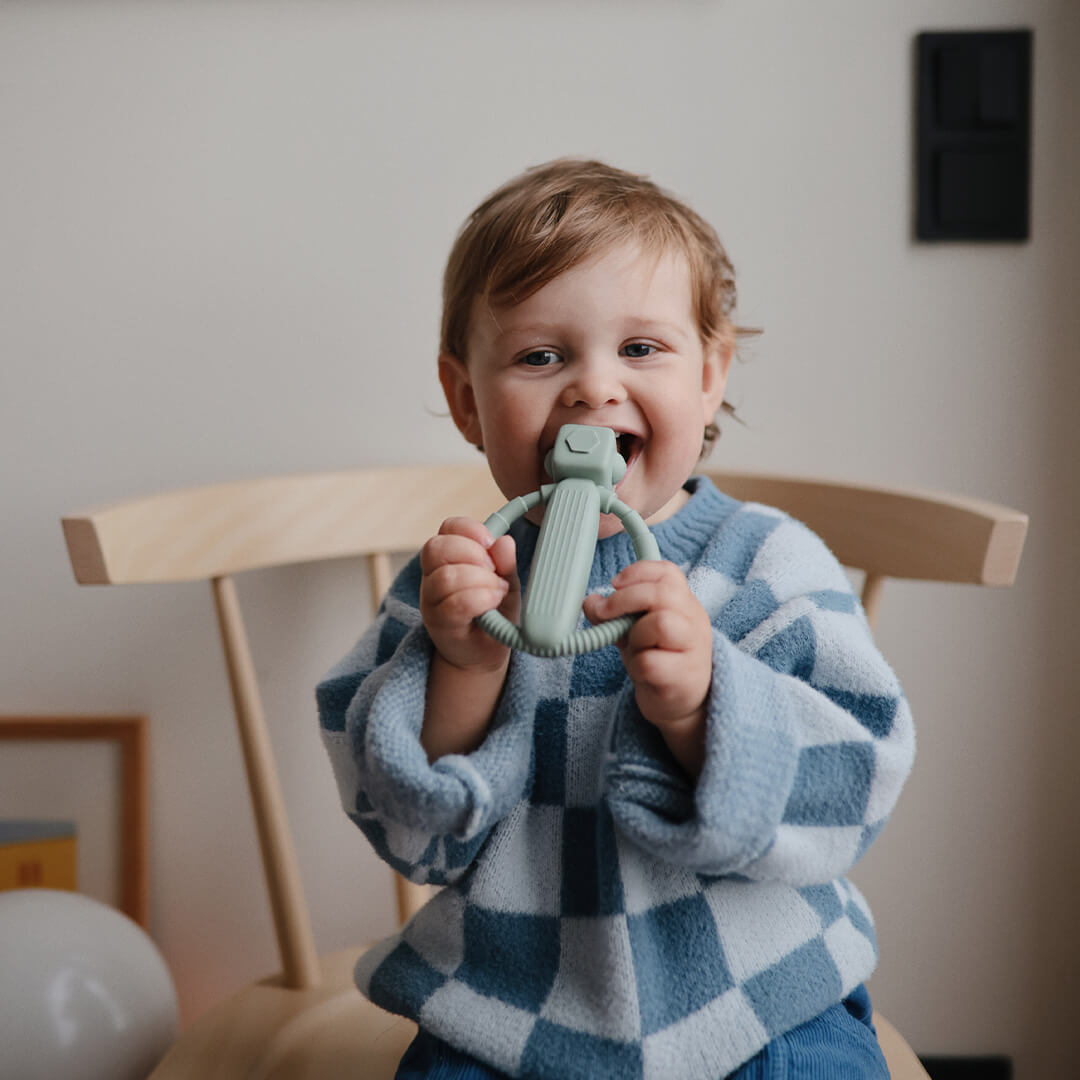 Een lachende peuter bijt op een mushie robot bijtspeelgoed terwijl hij op een houten stoel zit.