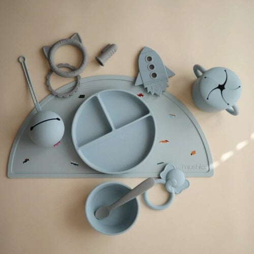 Een collectie mushie babyspullen, waaronder een blauwe raket bijtspeeltje, een placemat, bord, kom en andere siliconen speeltjes.