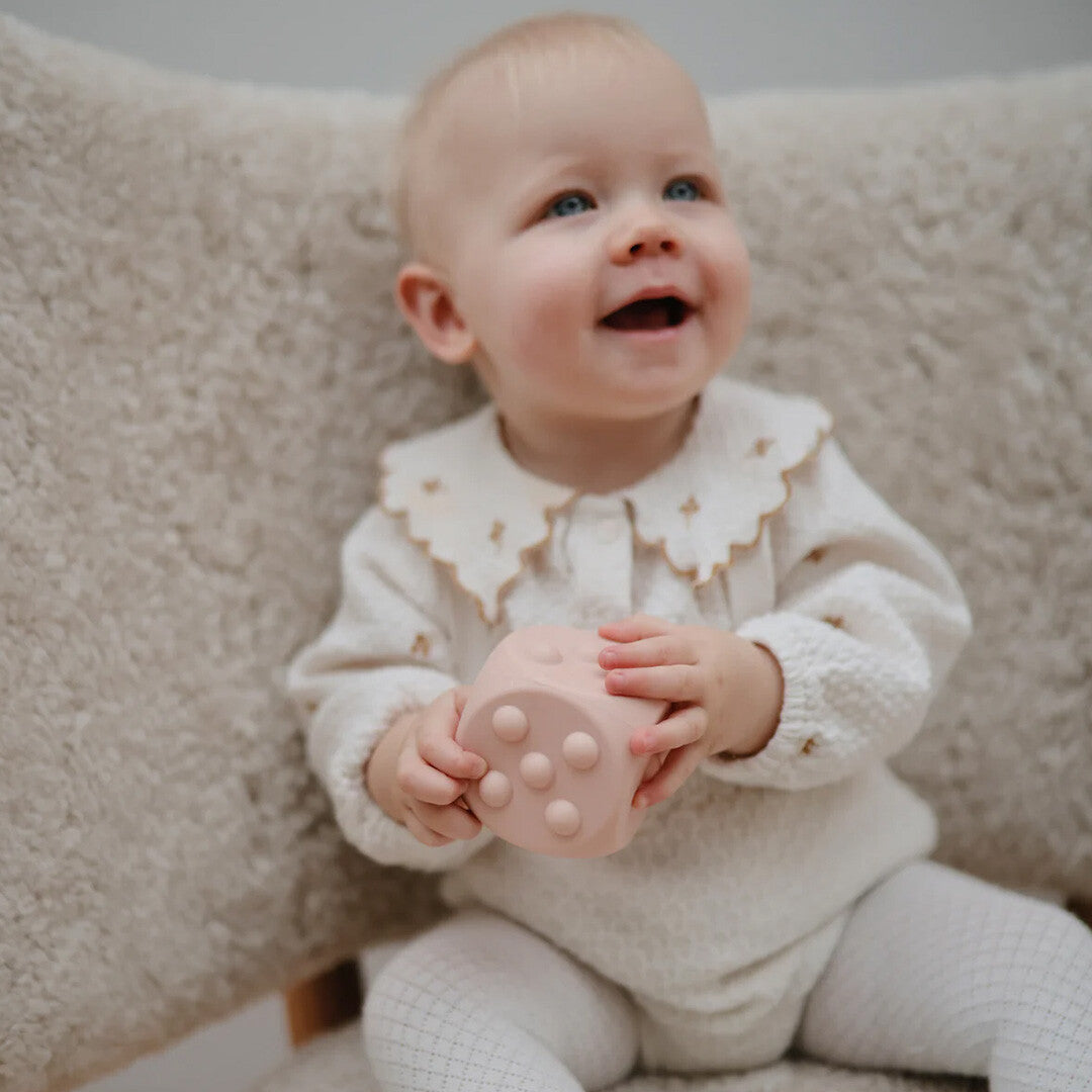 Een lachende baby met blauwe ogen houdt een blush roze mushie siliconen sensorische dobbelsteen vast.