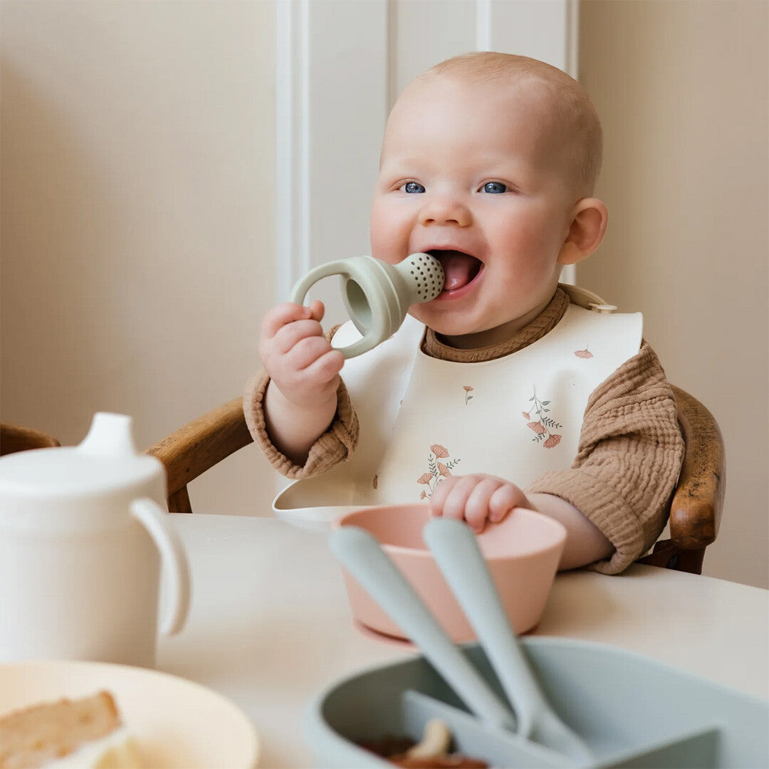 Een baby in een kinderstoel die blij op een mushie fruitspeen kauwt.