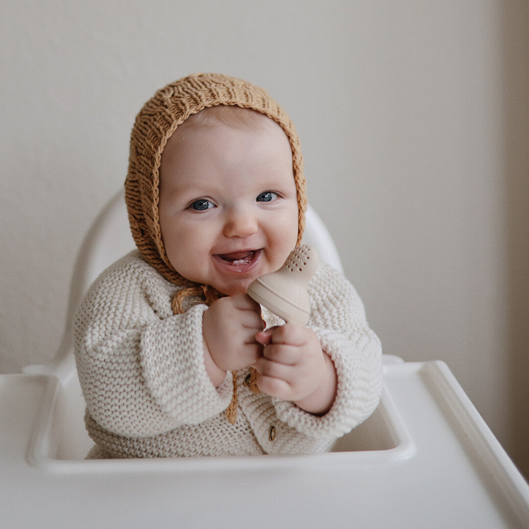 Een lachende baby in een kinderstoel houdt een beige Mushie fruitspeen vast.