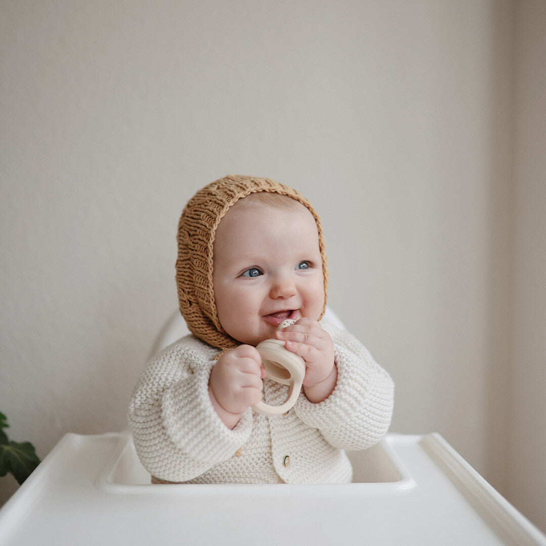 Een blije baby in een kinderstoel kijkt opzij terwijl het een beige Mushie fruitspeen vasthoudt.
