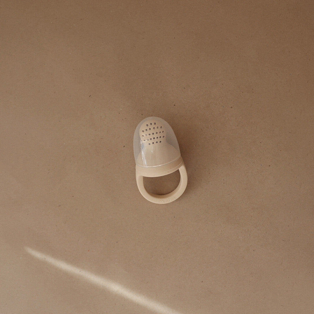 Een beige Mushie fruitspeen met transparante dop ligt op een bruine ondergrond.