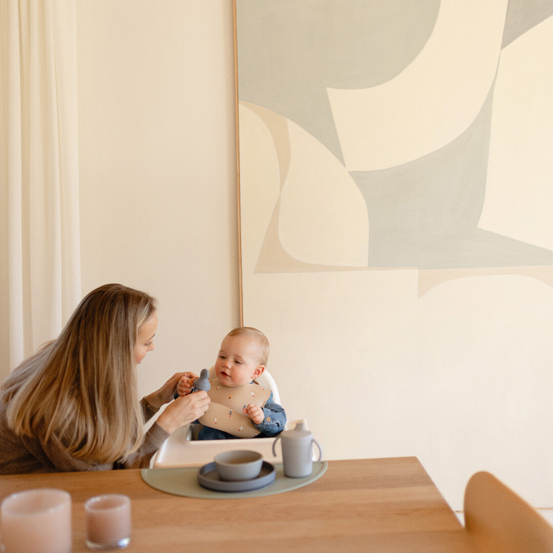Een moeder geeft een baby in een kinderstoel een mushie fruitspeen.
