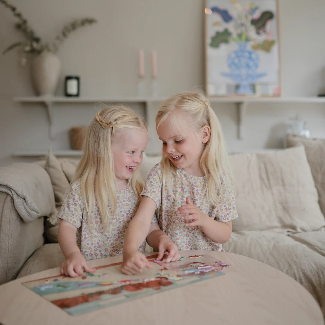 Twee lachende meisjes spelen samen met de Mushie herbruikbare dinosaurussticker set aan een houten tafel.