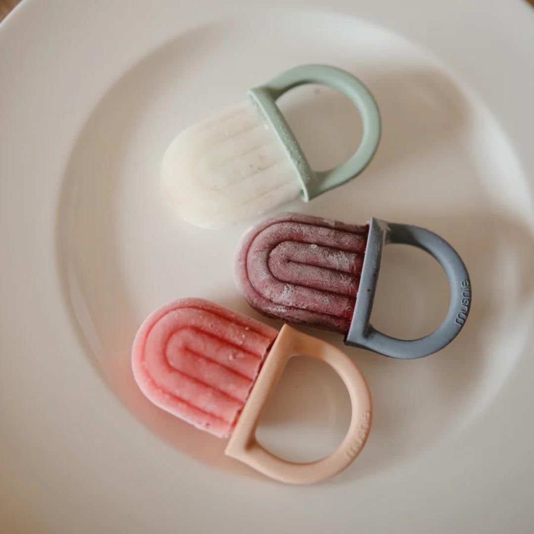 Drie Mushie ijsvormen met fruitijsjes in wit, paars en roze op een wit bord.