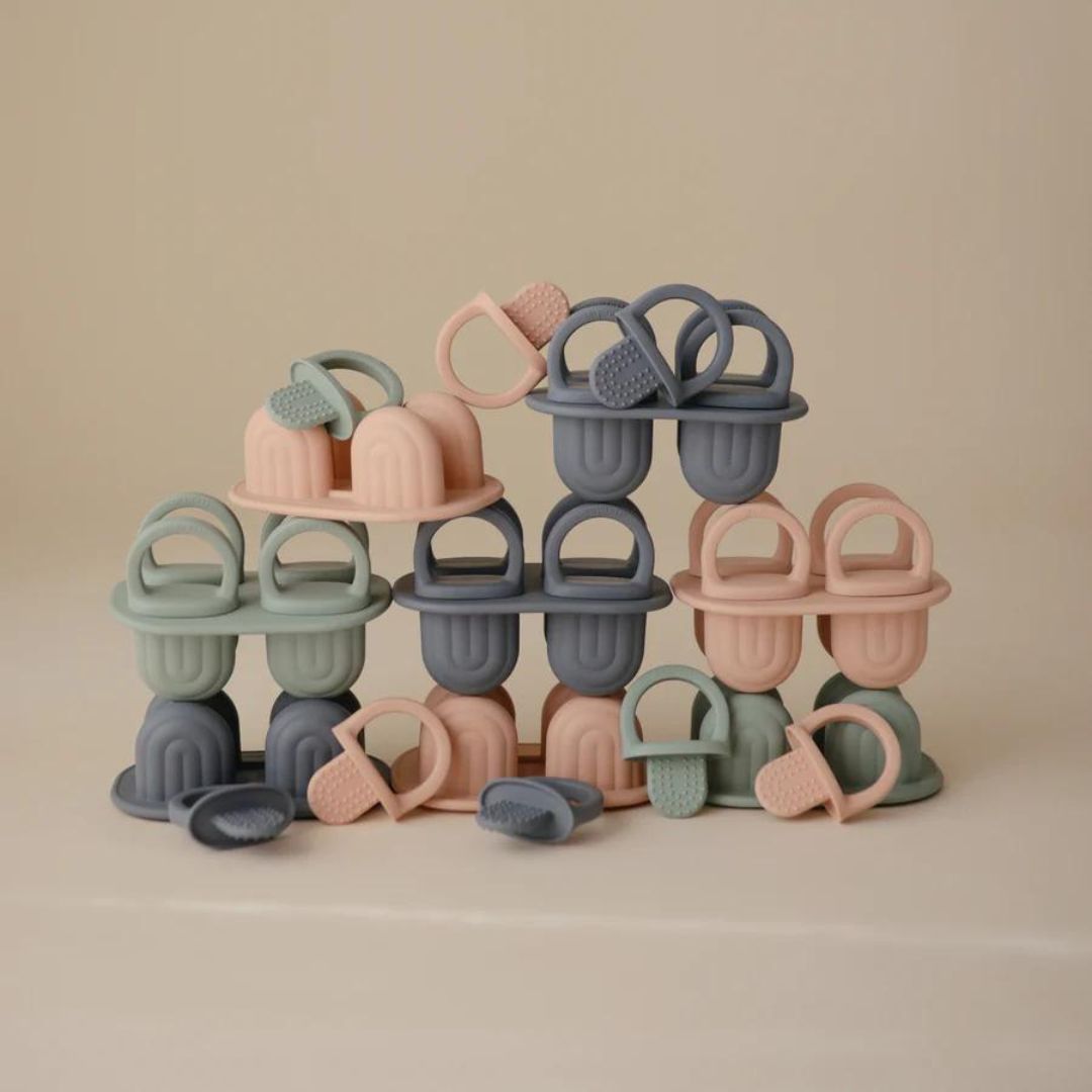 Een stapel Mushie siliconen ijsvormen in pastelkleuren blauw, roze en groen, met losse bijtringen.