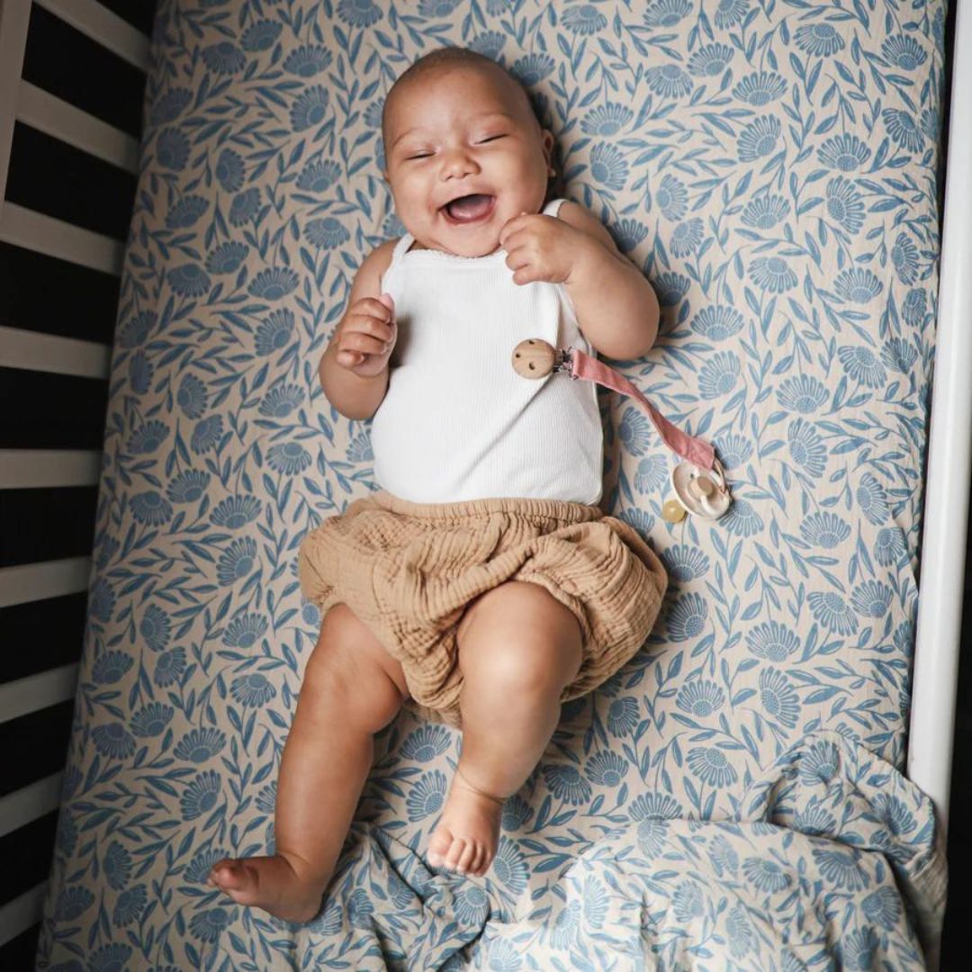Lachende baby ligt in een wieg op de mushie inbakerdoek met blauw bloemenpatroon.
