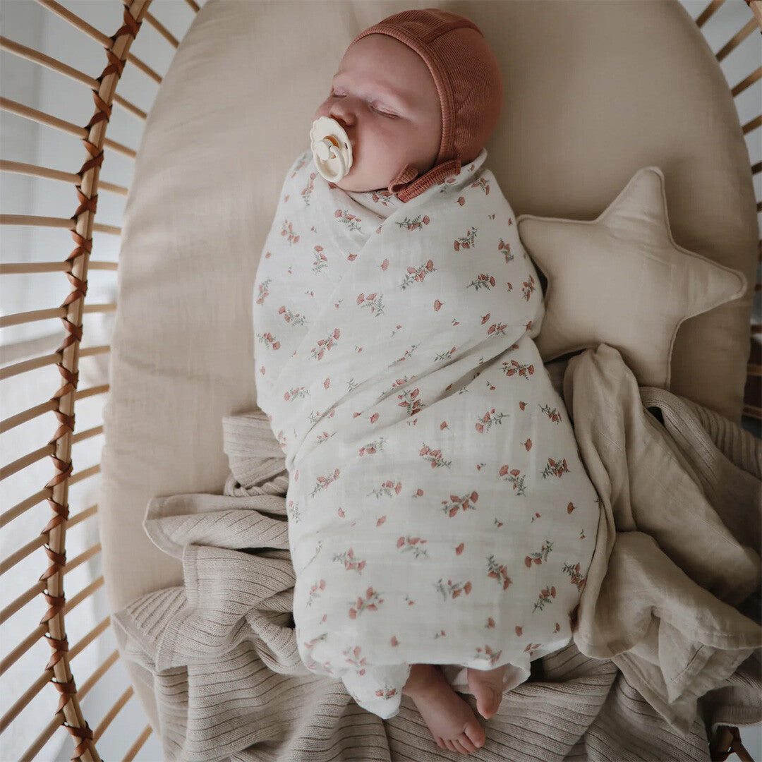 Slapende baby ingebakerd in een mushie inbakerdoek met roze bloemenpatroon in een wieg.