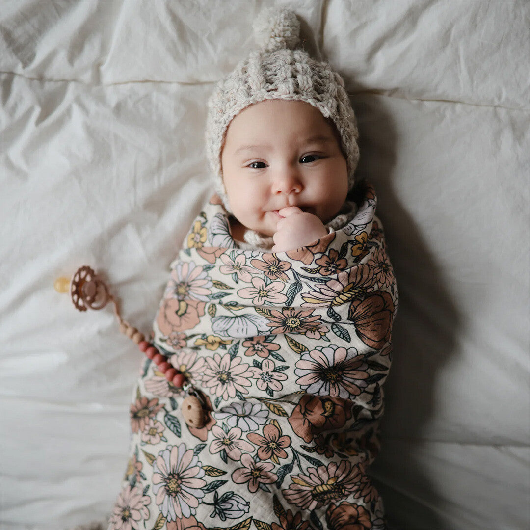 Een baby ingebakerd in een mushie inbakerdoek met retro bloemenpatroon en een gebreide muts.