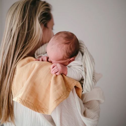 Een baby slaapt op de schouder van een volwassene, deels bedekt met een gestreepte mushie inbakerdoek.