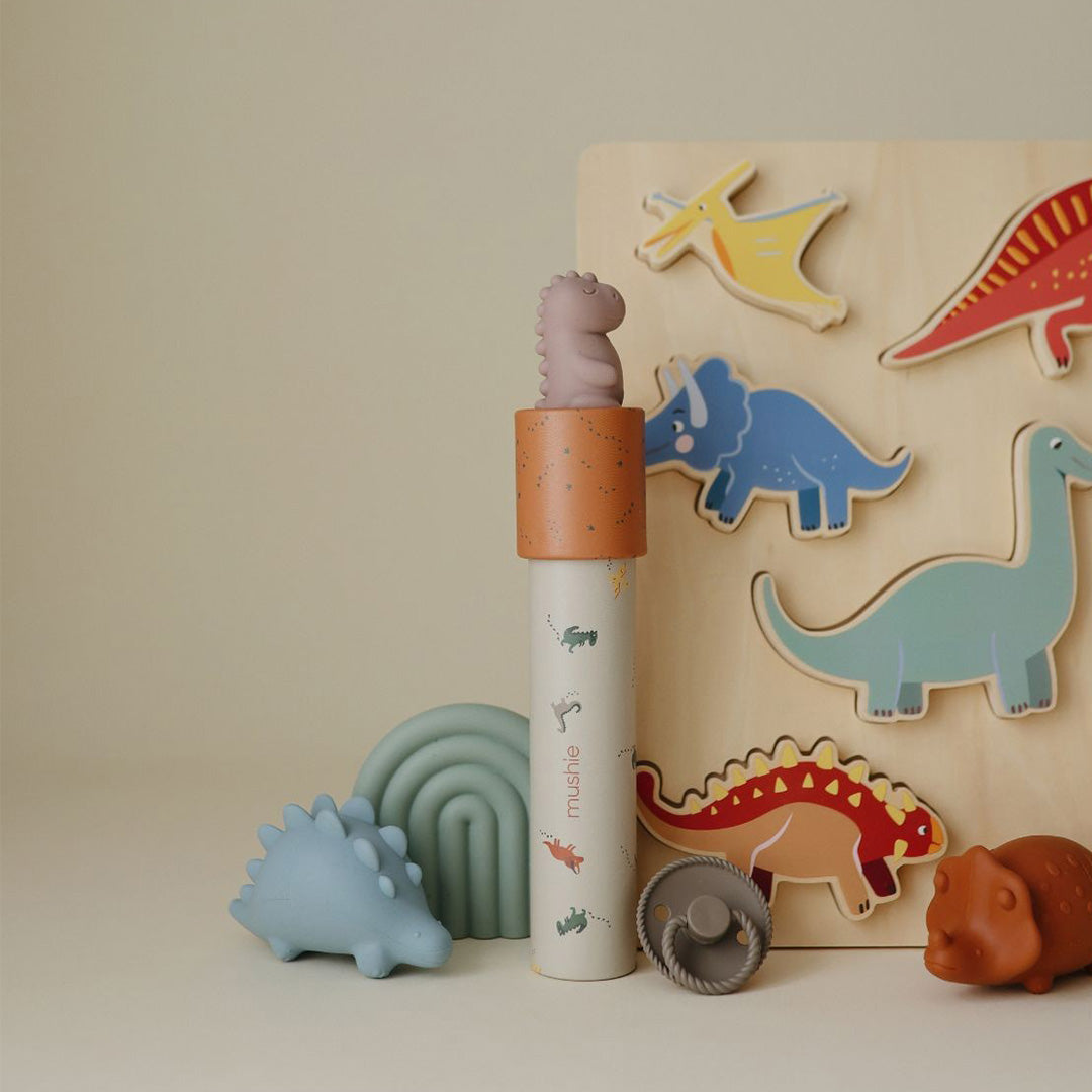 Een Mushie kaleidoscoop met dinosaurus-thema staat rechtop tussen diverse dinosaurus-speeltjes.