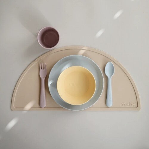 Een enkele gedekte plaats met een mushie placemat, bord, kom, vork en lepel in pastelkleuren.